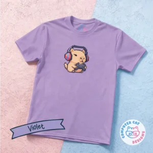A violet-colored t-shirt on a pink and blue surface featuring a pixelated graphic of a capybara playing videogames, with a controller in it's paws and red headphones on. The shirt has a Computer Cat Designs logo.